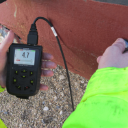 Person in a bright neon jacket uses an ultrasonic thickness gauge on a red surface, showing a reading of 4.7 on the screen.