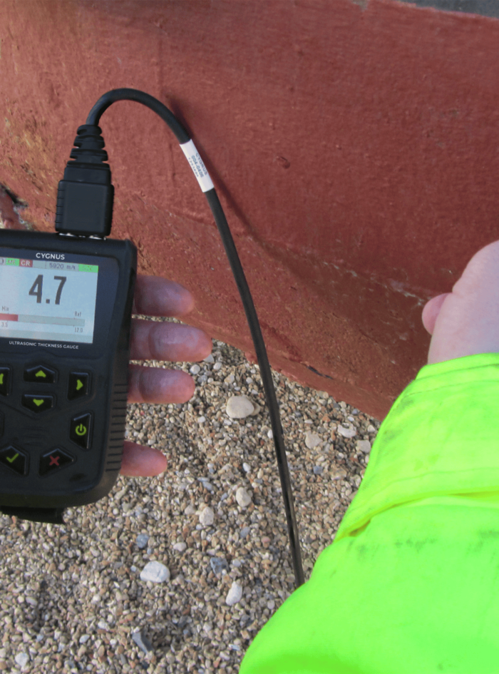 Person in a bright neon jacket uses an ultrasonic thickness gauge on a red surface, showing a reading of 4.7 on the screen.